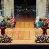 El secretario de Estado de EEUU, Marco Rubio (traje azul marino, camisa blanca y corbata azul marino con puntos blancos), y el presidente de Guatemala, Bernardo Arévalo (traje gris oscuro, camisa blanca y corbata azul cielo), durante una rueda de prensa conjunta en el Palacio Nacional de la Cultura (Ciudad de Guatemala), el 5 de febrero de 2025. Ambos están de pie detrás de podios de madera con el escudo nacional de Guatemala, sobre una tarima elevada con arreglos florales al frente. A sus lados se encuentran las banderas de Estados Unidos y Guatemala sobre soportes. De fondo, se aprecia la decoración del espacio, con arcos ornamentados y vegetación. Marco Rubio