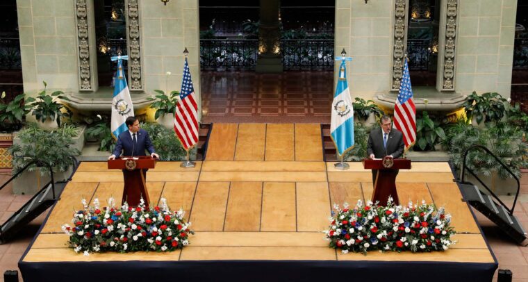 El secretario de Estado de EEUU, Marco Rubio (traje azul marino, camisa blanca y corbata azul marino con puntos blancos), y el presidente de Guatemala, Bernardo Arévalo (traje gris oscuro, camisa blanca y corbata azul cielo), durante una rueda de prensa conjunta en el Palacio Nacional de la Cultura (Ciudad de Guatemala), el 5 de febrero de 2025. Ambos están de pie detrás de podios de madera con el escudo nacional de Guatemala, sobre una tarima elevada con arreglos florales al frente. A sus lados se encuentran las banderas de Estados Unidos y Guatemala sobre soportes. De fondo, se aprecia la decoración del espacio, con arcos ornamentados y vegetación. Marco Rubio