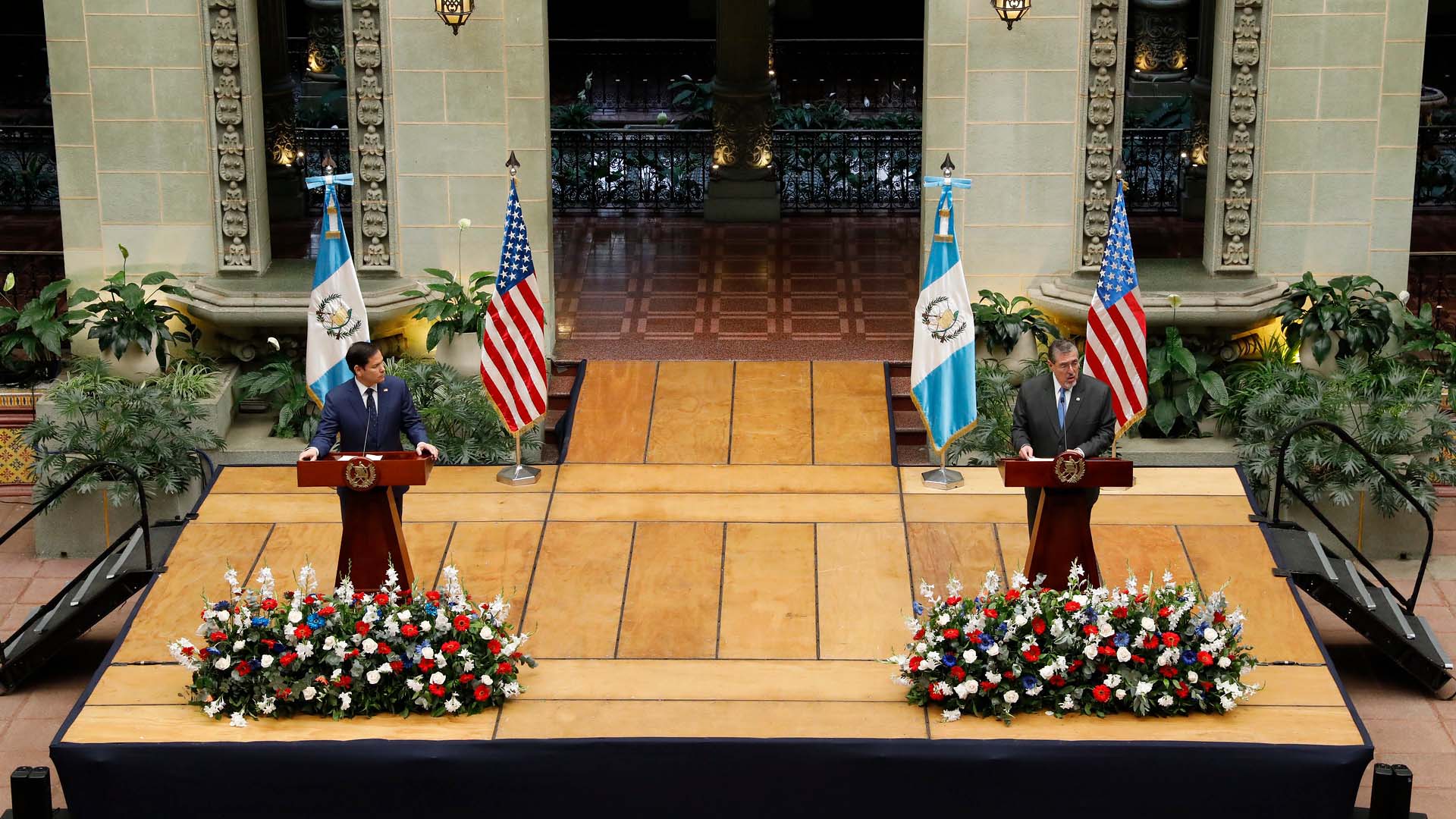 El secretario de Estado de EEUU, Marco Rubio (traje azul marino, camisa blanca y corbata azul marino con puntos blancos), y el presidente de Guatemala, Bernardo Arévalo (traje gris oscuro, camisa blanca y corbata azul cielo), durante una rueda de prensa conjunta en el Palacio Nacional de la Cultura (Ciudad de Guatemala), el 5 de febrero de 2025. Ambos están de pie detrás de podios de madera con el escudo nacional de Guatemala, sobre una tarima elevada con arreglos florales al frente. A sus lados se encuentran las banderas de Estados Unidos y Guatemala sobre soportes. De fondo, se aprecia la decoración del espacio, con arcos ornamentados y vegetación. Marco Rubio