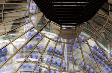 Primer plano de la estructura de la cúpula del edificio Reichstag en Berlín. En la parte superior, se aprecia el cimborrio de la cúpula, con detalles metálicos oscuros y el vidrio que completa su estructura. En el centro de la imagen, los reflejos en los paneles multiplican la vista de la sala de plenos del edificio, situada en el fondo. Esta sala tiene una disposición semicircular con asientos de color azul, organizados alrededor de un espacio central. Alemania