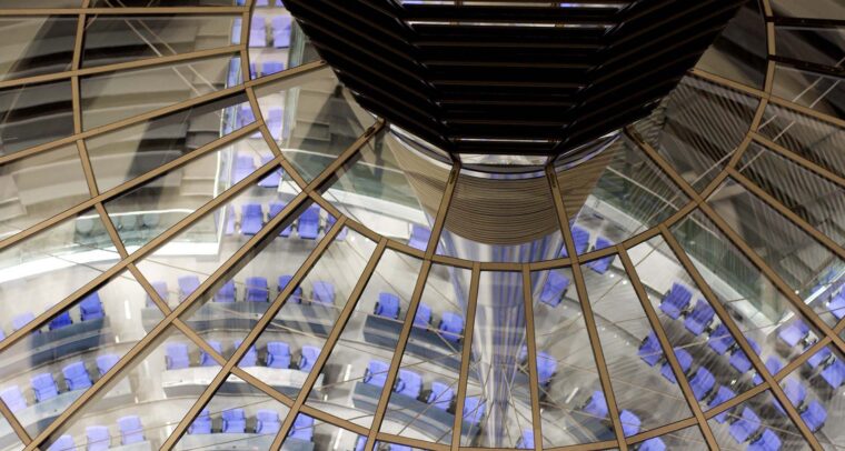 Primer plano de la estructura de la cúpula del edificio Reichstag en Berlín. En la parte superior, se aprecia el cimborrio de la cúpula, con detalles metálicos oscuros y el vidrio que completa su estructura. En el centro de la imagen, los reflejos en los paneles multiplican la vista de la sala de plenos del edificio, situada en el fondo. Esta sala tiene una disposición semicircular con asientos de color azul, organizados alrededor de un espacio central. Alemania