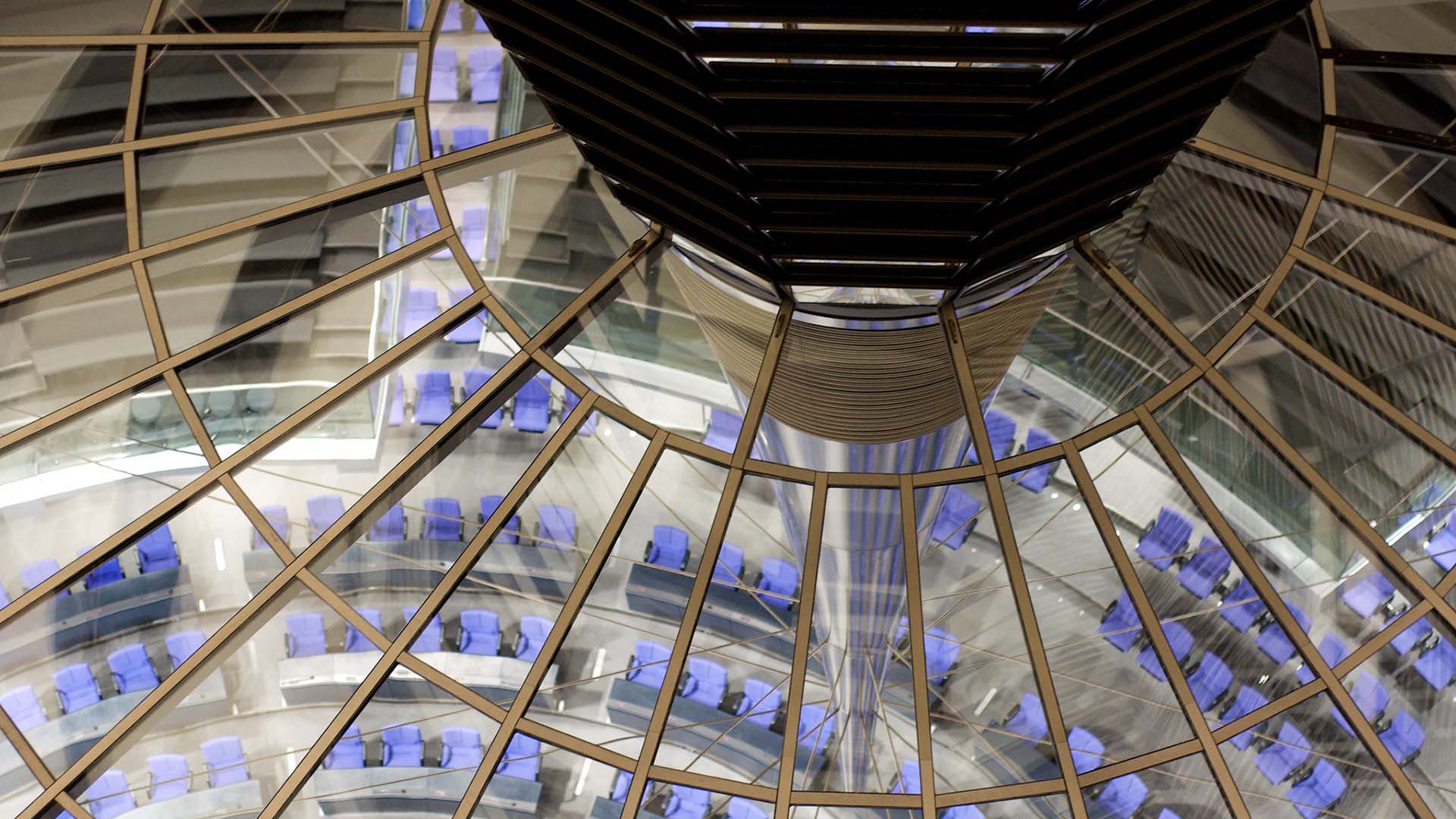 Primer plano de la estructura de la cúpula del edificio Reichstag en Berlín. En la parte superior, se aprecia el cimborrio de la cúpula, con detalles metálicos oscuros y el vidrio que completa su estructura. En el centro de la imagen, los reflejos en los paneles multiplican la vista de la sala de plenos del edificio, situada en el fondo. Esta sala tiene una disposición semicircular con asientos de color azul, organizados alrededor de un espacio central. Alemania