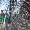 Primer plano a través de un alambre de púas gris enmarañado, colocado en el muro fronterizo oeste del Puerto de San Luis, en Arizona (Estados Unidos). En el fondo, un montacargas verde sostiene una plataforma elevada, sobre la cual dos marines del ejército estadounidense trabajan en la instalación del alambre. También se distingue la valla fronteriza de color marrón bajo un cielo despejado y soleado. Trump