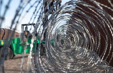 Primer plano a través de un alambre de púas gris enmarañado, colocado en el muro fronterizo oeste del Puerto de San Luis, en Arizona (Estados Unidos). En el fondo, un montacargas verde sostiene una plataforma elevada, sobre la cual dos marines del ejército estadounidense trabajan en la instalación del alambre. También se distingue la valla fronteriza de color marrón bajo un cielo despejado y soleado. Trump