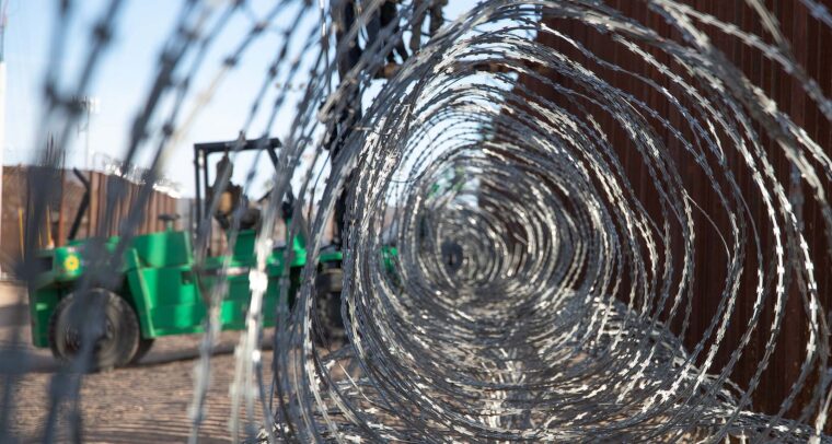 Primer plano a través de un alambre de púas gris enmarañado, colocado en el muro fronterizo oeste del Puerto de San Luis, en Arizona (Estados Unidos). En el fondo, un montacargas verde sostiene una plataforma elevada, sobre la cual dos marines del ejército estadounidense trabajan en la instalación del alambre. También se distingue la valla fronteriza de color marrón bajo un cielo despejado y soleado. Trump