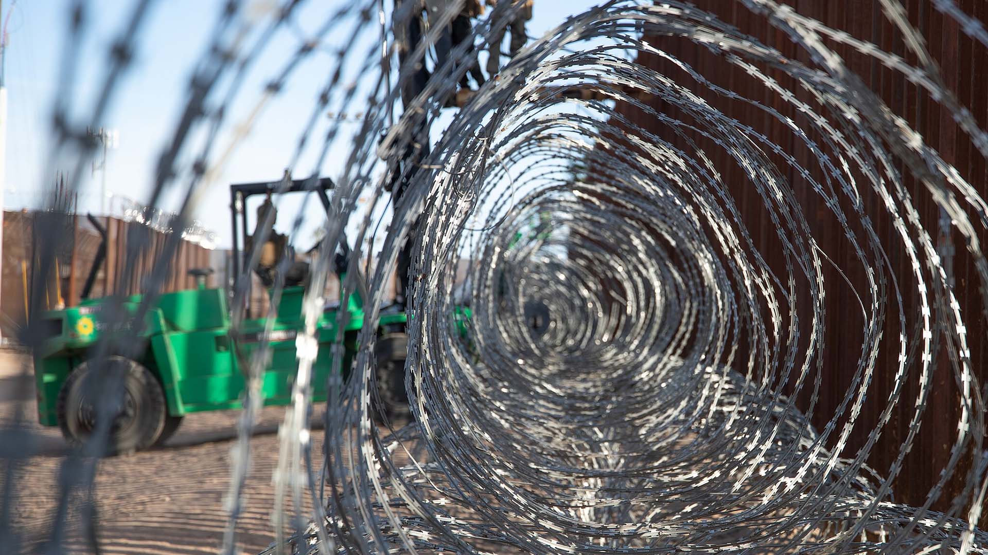 Close-up view through tangled grey barbed wire placed on the western border wall of the San Luis Port of Entry in Arizona (United States). In the background, a green forklift holds an elevated platform, on which two US Marines are working to install the wire. The brown border fence is also visible under a clear and sunny sky. Trump