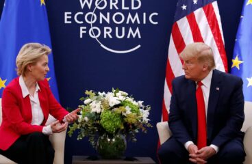 Ursula von der Leyen (chaqueta roja, camisa blanca y pantalón negro), presidenta de la Comisión Europea, y Donald Trump (traje azul marino, corbata roja y camisa blanca), presidente de Estados Unidos, se reúnen durante el Foro Económico Mundial en Davos en 2020. Ambos están sentados en unos sillones color crema, con una mesa de café entre ellos, presidida por un arreglo de flores blancas y verdes. Detrás, sobre un fondo azul oscuro, se lee ‘World Economic Forum’, con las banderas de la UE y de Estados Unidos situadas a los lados. Aranceles de Trump