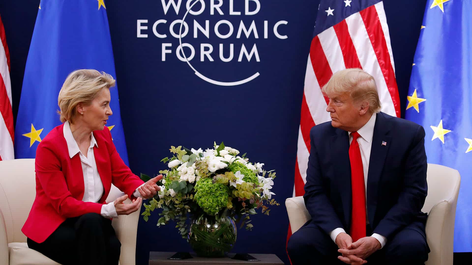 Ursula von der Leyen (chaqueta roja, camisa blanca y pantalón negro), presidenta de la Comisión Europea, y Donald Trump (traje azul marino, corbata roja y camisa blanca), presidente de Estados Unidos, se reúnen durante el Foro Económico Mundial en Davos en 2020. Ambos están sentados en unos sillones color crema, con una mesa de café entre ellos, presidida por un arreglo de flores blancas y verdes. Detrás, sobre un fondo azul oscuro, se lee ‘World Economic Forum’, con las banderas de la UE y de Estados Unidos situadas a los lados. Aranceles de Trump