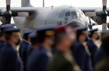 Acto oficial de despedida del avión de transporte militar T.10 en la base aérea de Zaragoza. En el fondo, resaltado, se encuentra el fuselaje del avión de color gris, con algunos detalles en amarillo. Tiene alas altas y dos grandes hélices visibles, una a cada lado. En la parte delantera, destacan los cristales inclinados de la cabina y el número 07. En la parte delantera de la imagen, desenfocadas, varias personas del Ejército del Aire están en formación. La mayoría viste uniforme azul oscuro con gorras del mismo color. Destaca una persona en el centro con un uniforme militar verde. Defensa