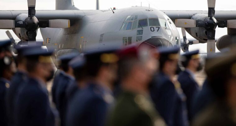 Official farewell ceremony for the T.10 military transport aircraft at Zaragoza Air Base. In the background, prominently displayed, is the grey fuselage of the aircraft, with some yellow details. It has high wings and two large visible propellers, one on each side. At the front, the slanted cockpit windows and the number 07 stand out. In the foreground of the image, blurred, several members of the Air Force are in formation. Most are wearing dark blue uniforms with matching caps. A person in the centre stands out, dressed in a green military uniform. Defence