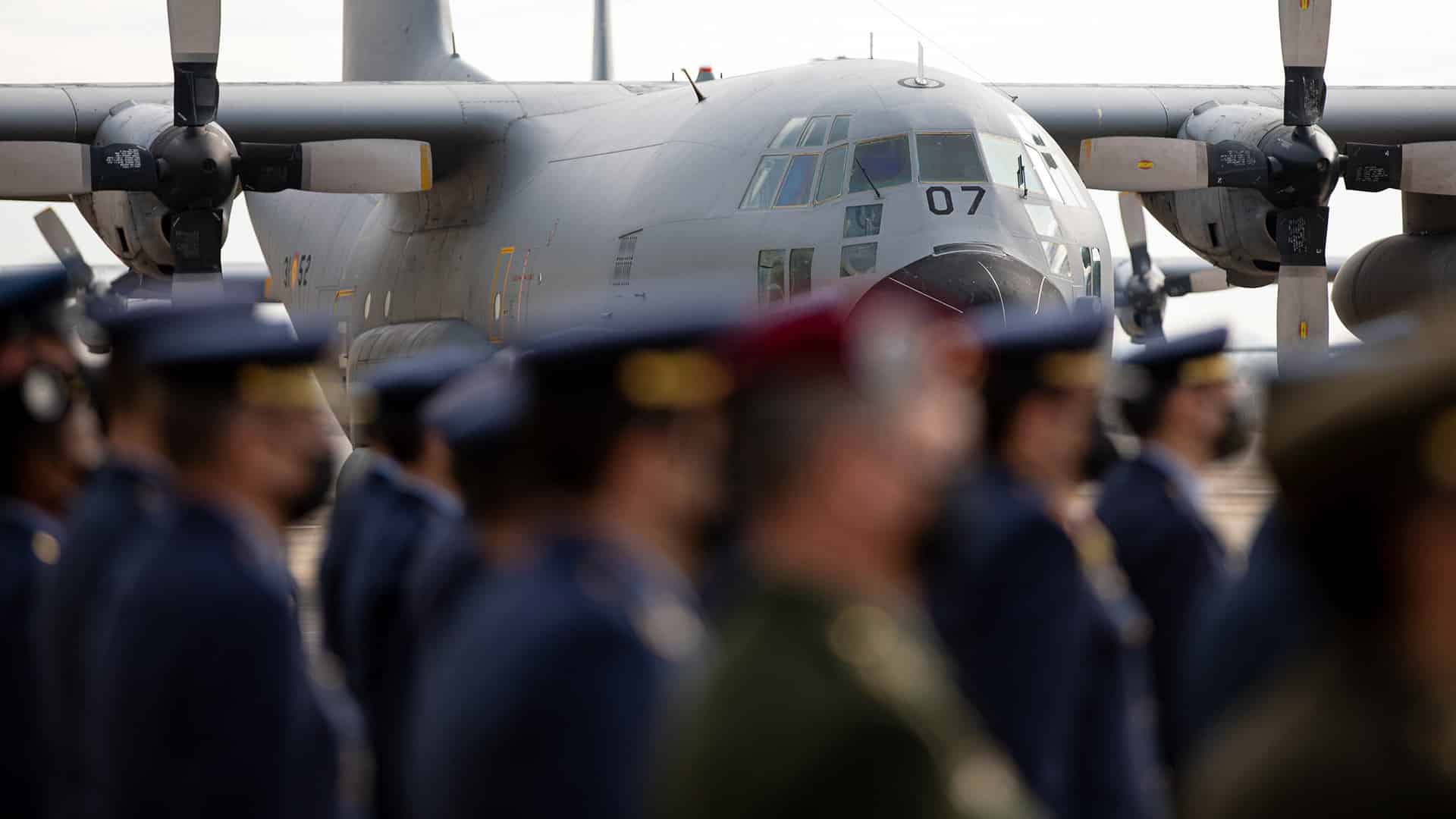 Acto oficial de despedida del avión de transporte militar T.10 en la base aérea de Zaragoza. En el fondo, resaltado, se encuentra el fuselaje del avión de color gris, con algunos detalles en amarillo. Tiene alas altas y dos grandes hélices visibles, una a cada lado. En la parte delantera, destacan los cristales inclinados de la cabina y el número 07. En la parte delantera de la imagen, desenfocadas, varias personas del Ejército del Aire están en formación. La mayoría viste uniforme azul oscuro con gorras del mismo color. Destaca una persona en el centro con un uniforme militar verde. Defensa