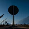 View of the Gemasolar solar power plant in Fuentes de Andalucía (Spain), featuring large reflective panels and heliostat structures tilted towards the sky. In the centre of the image, a circular traffic sign partially obstructs the view of the technological landscape. Just transition