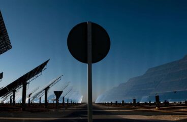 View of the Gemasolar solar power plant in Fuentes de Andalucía (Spain), featuring large reflective panels and heliostat structures tilted towards the sky. In the centre of the image, a circular traffic sign partially obstructs the view of the technological landscape. Just transition