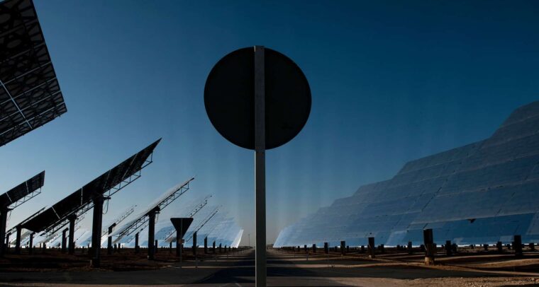 View of the Gemasolar solar power plant in Fuentes de Andalucía (Spain), featuring large reflective panels and heliostat structures tilted towards the sky. In the centre of the image, a circular traffic sign partially obstructs the view of the technological landscape. Just transition