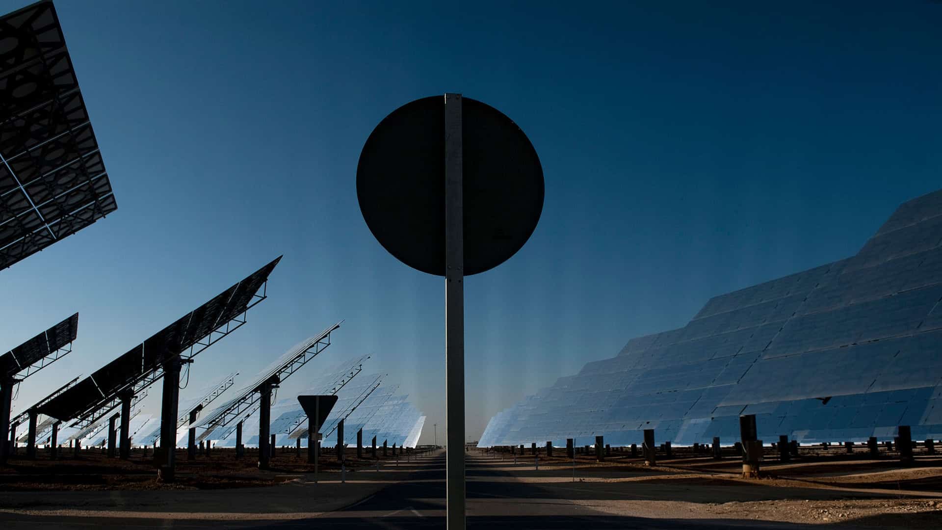 View of the Gemasolar solar power plant in Fuentes de Andalucía (Spain), featuring large reflective panels and heliostat structures tilted towards the sky. In the centre of the image, a circular traffic sign partially obstructs the view of the technological landscape. Just transition