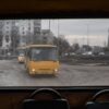 A view from inside an old yellow bus shows a war-torn cityscape in Borodyanka, Ukraine. Through the rear window, two yellow buses are driving along a muddy road, passing a heavily damaged block of flats. In the background, a monument with a Ukrainian flag on top can be seen. Ukraine