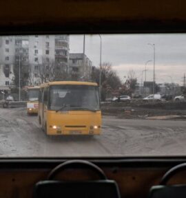 Vista desde el interior de un antiguo autobús amarillo que muestra un paisaje urbano devastado por la guerra en Borodyanka, Ucrania. A través de la ventana trasera, dos autobuses amarillos circulan por un camino embarrado, pasando junto a un edificio de apartamentos gravemente dañado. En el fondo, se aprecia monumento con una bandera ucraniana en la cima. Ucrania