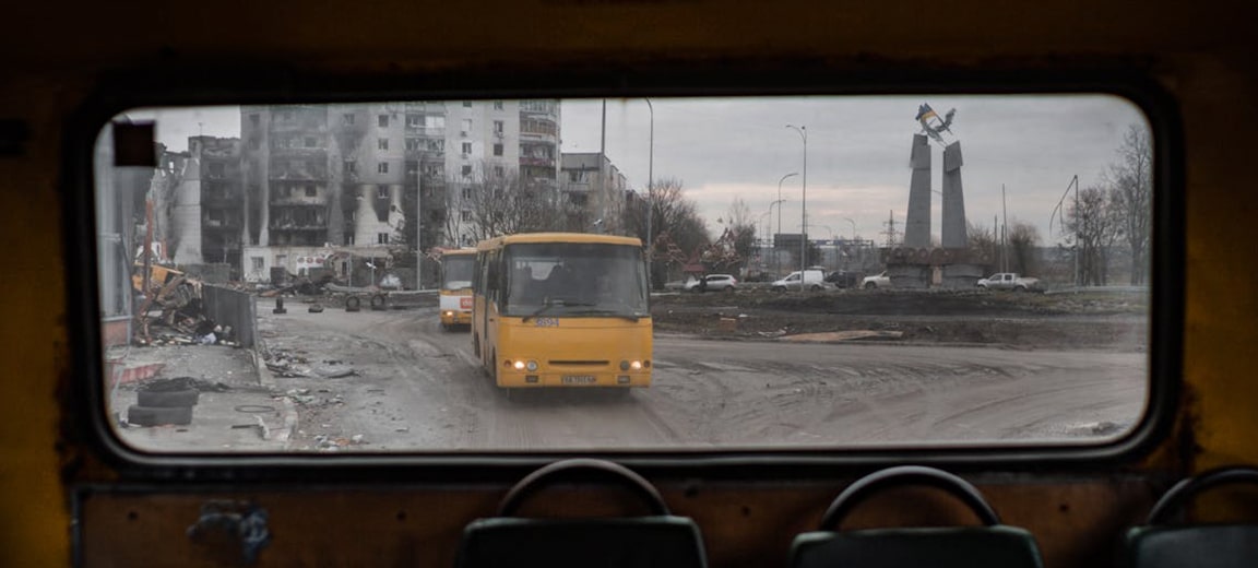 A view from inside an old yellow bus shows a war-torn cityscape in Borodyanka, Ukraine. Through the rear window, two yellow buses are driving along a muddy road, passing a heavily damaged block of flats. In the background, a monument with a Ukrainian flag on top can be seen. Ukraine