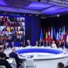 Detalle de la cumbre internacional en Kyiv, presidida por Ursula von der Leyen, con líderes europeos e internacionales reunidos en una mesa circular para debatir el apoyo a Ucrania en el tercer aniversario de la invasión rusa. A la izquierda de la sala, se ve proyectada en una pantalla grande la participación virtual de 20 participantes. A la derecha, una amalgama de banderas de diferentes regiones del mundo está colocada detrás de la mesa circular blanca donde los líderes están sentados. Se aprecian varias filas de sillas blancas alrededor de la mesa. UE