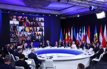 Detalle de la cumbre internacional en Kyiv, presidida por Ursula von der Leyen, con líderes europeos e internacionales reunidos en una mesa circular para debatir el apoyo a Ucrania en el tercer aniversario de la invasión rusa. A la izquierda de la sala, se ve proyectada en una pantalla grande la participación virtual de 20 participantes. A la derecha, una amalgama de banderas de diferentes regiones del mundo está colocada detrás de la mesa circular blanca donde los líderes están sentados. Se aprecian varias filas de sillas blancas alrededor de la mesa. UE