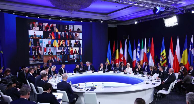 Detalle de la cumbre internacional en Kyiv, presidida por Ursula von der Leyen, con líderes europeos e internacionales reunidos en una mesa circular para debatir el apoyo a Ucrania en el tercer aniversario de la invasión rusa. A la izquierda de la sala, se ve proyectada en una pantalla grande la participación virtual de 20 participantes. A la derecha, una amalgama de banderas de diferentes regiones del mundo está colocada detrás de la mesa circular blanca donde los líderes están sentados. Se aprecian varias filas de sillas blancas alrededor de la mesa. UE