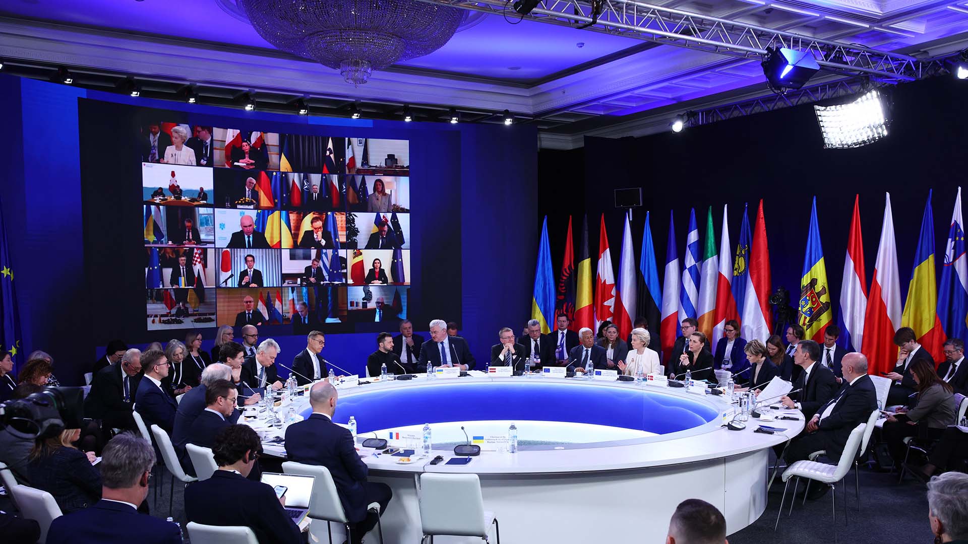 Detalle de la cumbre internacional en Kyiv, presidida por Ursula von der Leyen, con líderes europeos e internacionales reunidos en una mesa circular para debatir el apoyo a Ucrania en el tercer aniversario de la invasión rusa. A la izquierda de la sala, se ve proyectada en una pantalla grande la participación virtual de 20 participantes. A la derecha, una amalgama de banderas de diferentes regiones del mundo está colocada detrás de la mesa circular blanca donde los líderes están sentados. Se aprecian varias filas de sillas blancas alrededor de la mesa. UE