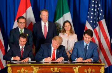 De izquierda a derecha, sentados y firmando el tratado de libre comercio entre EEUU, Canadá y México (USMCA) en la cumbre del G20 en Argentina en el año 2018: Enrique Peña Nieto, presidente de México, con un traje negro, camisa blanca y corbata a rayas rojas y azules; Donald Trump, presidente de EEUU, con un traje azul oscuro, camisa blanca y corbata roja; y Justin Trudeau, primer ministro de Canadá, con un traje, camisa y corbata en distintas tonalidades de azul. Al fondo, las banderas de Estados Unidos, Canadá y México, así como tres personas que acompañan la firma (dos hombres y una mujer de pie), con un fondo azul.