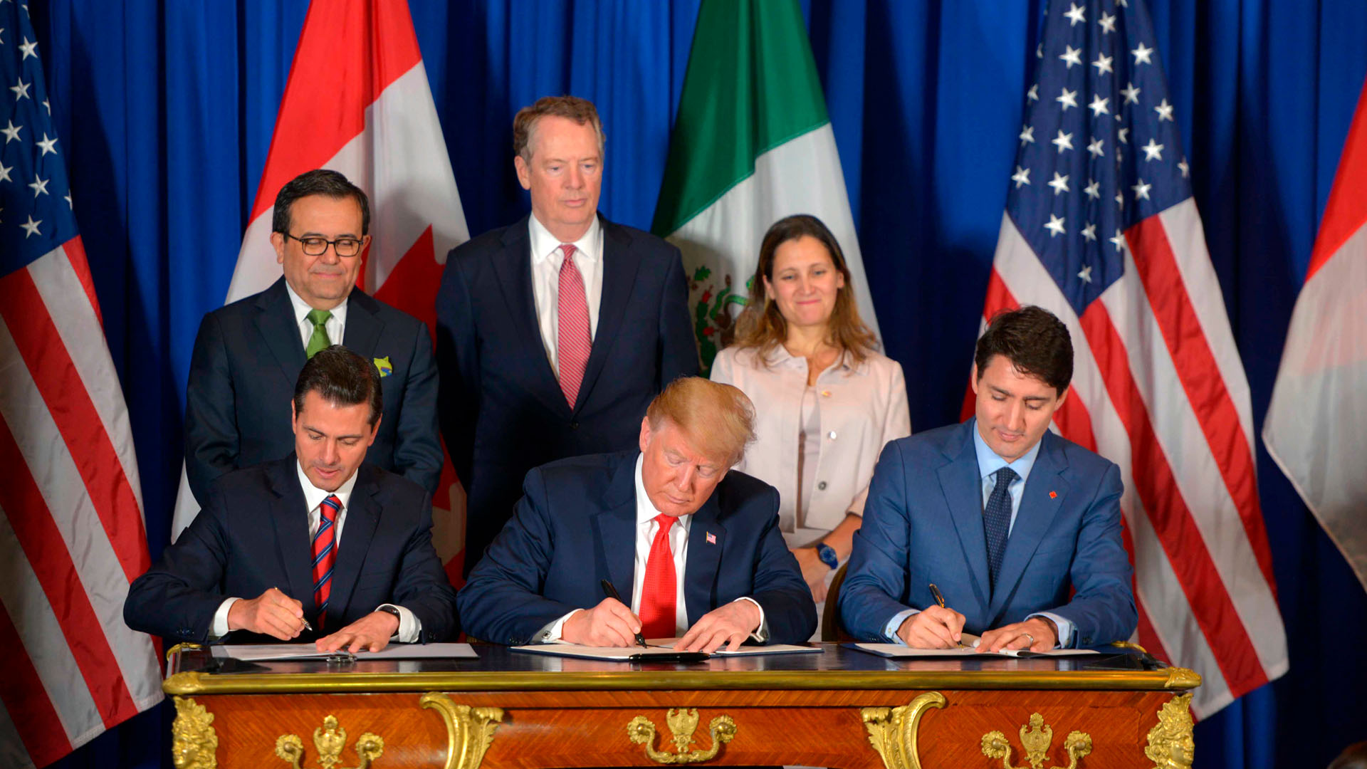 En primer plano, de izquierda a derecha, se encuentran sentados y firmando el tratado de libre comercio entre EEUU, Canadá y México (USMCA) en la cumbre del G20 en Argentina en el año 2018: Enrique Peña Nieto, expresidente de México, con un traje negro, camisa blanca y corbata a rayas rojas y azules; Donald Trump, presidente de EEUU, con un traje azul oscuro, camisa blanca y corbata roja; y Justin Trudeau, primer ministro de Canadá, con un traje, camisa y corbata en distintas tonalidades de azul. Al fondo, se vislumbran las banderas de Estados Unidos, Canadá y México, así como tres personas que acompañan la firma (dos hombres y una mujer de pie), con un fondo azul. Trump