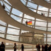 Interior de la cúpula del Reichstag en Berlín durante el día. En primer plano, se aprecia su estructura circular de varias plantas, de color blanco, con barandillas acristaladas. Detrás, se observa la estructura de la cúpula con los mismos detalles y una parte del edificio, sobre el que ondea la bandera alemana, al fondo. En la parte inferior, se ven varias personas caminando o contemplando detalles de la estructura. Alemania