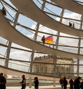 Interior de la cúpula del Reichstag en Berlín durante el día. En primer plano, se aprecia su estructura circular de varias plantas, de color blanco, con barandillas acristaladas. Detrás, se observa la estructura de la cúpula con los mismos detalles y una parte del edificio, sobre el que ondea la bandera alemana, al fondo. En la parte inferior, se ven varias personas caminando o contemplando detalles de la estructura. Alemania