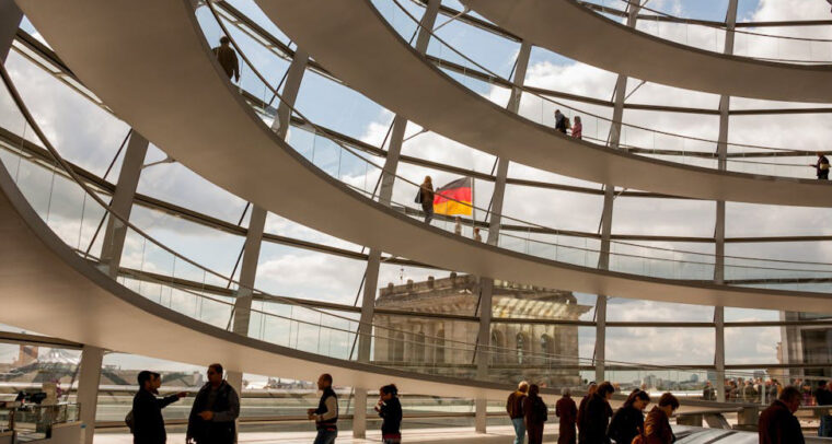 Interior de la cúpula del Reichstag en Berlín durante el día. En primer plano, se aprecia su estructura circular de varias plantas, de color blanco, con barandillas acristaladas. Detrás, se observa la estructura de la cúpula con los mismos detalles y una parte del edificio, sobre el que ondea la bandera alemana, al fondo. En la parte inferior, se ven varias personas caminando o contemplando detalles de la estructura. Alemania