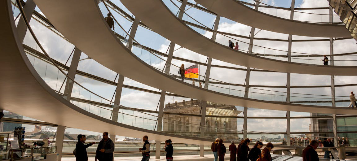 Interior de la cúpula del Reichstag en Berlín durante el día. En primer plano, se aprecia su estructura circular de varias plantas, de color blanco, con barandillas acristaladas. Detrás, se observa la estructura de la cúpula con los mismos detalles y una parte del edificio, sobre el que ondea la bandera alemana, al fondo. En la parte inferior, se ven varias personas caminando o contemplando detalles de la estructura. Alemania