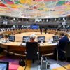 Roundtable discussion at the EU-Western Balkans Summit in Brussels on 18 December 2024, held in a modern conference hall with a multi-coloured geometric ceiling and a circular table. European and Western Balkans leaders are seated with laptops, microphones, and screens, engaging in discussions. A blue backdrop displays the event title, while glass windows and balconies accommodate translators and additional staff. Western Balkans