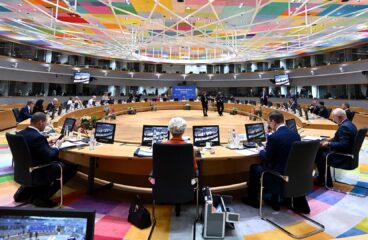 Roundtable discussion at the EU-Western Balkans Summit in Brussels on 18 December 2024, held in a modern conference hall with a multi-coloured geometric ceiling and a circular table. European and Western Balkans leaders are seated with laptops, microphones, and screens, engaging in discussions. A blue backdrop displays the event title, while glass windows and balconies accommodate translators and additional staff. Western Balkans