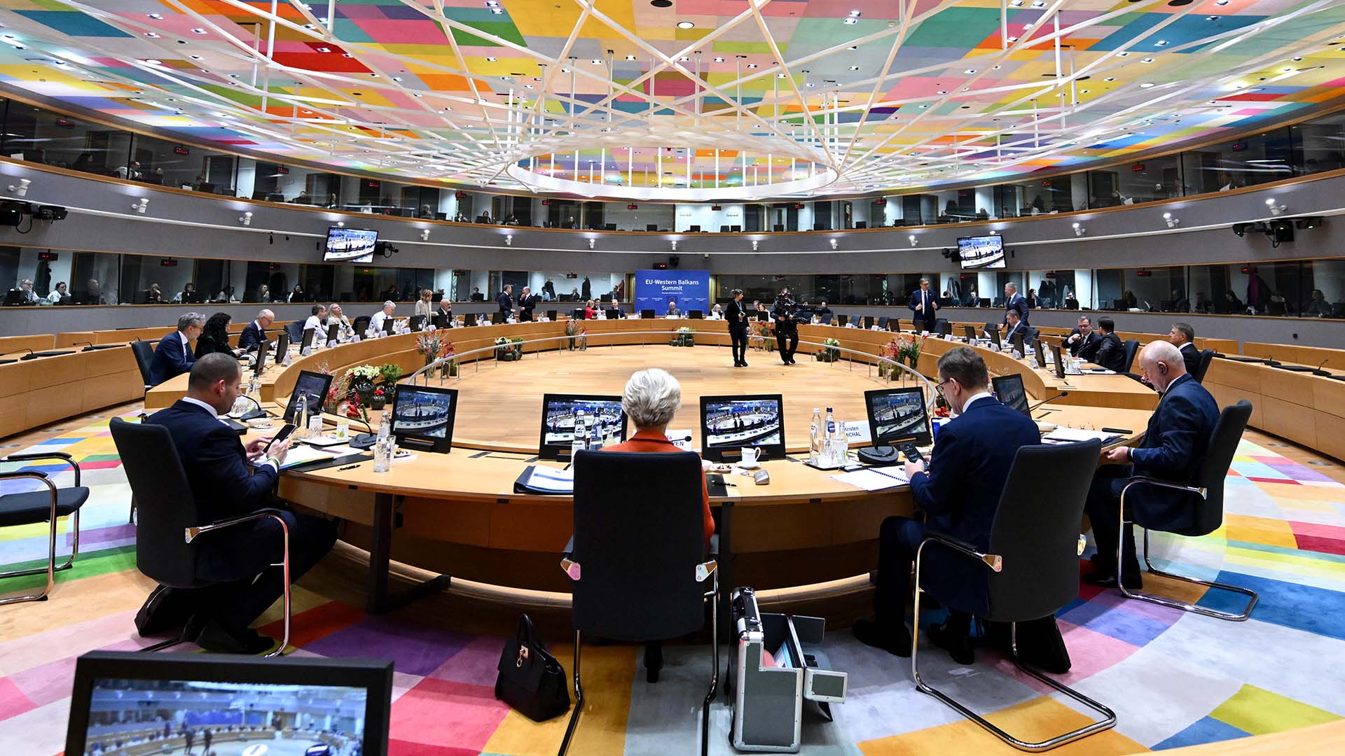 Roundtable discussion at the EU-Western Balkans Summit in Brussels on 18 December 2024, held in a modern conference hall with a multi-coloured geometric ceiling and a circular table. European and Western Balkans leaders are seated with laptops, microphones, and screens, engaging in discussions. A blue backdrop displays the event title, while glass windows and balconies accommodate translators and additional staff. Western Balkans