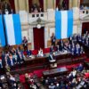 The President of Argentina, Javier Milei, stands at the centre of Congress, delivering a speech during the opening of the 2025 legislative session. Surrounding him, legislators, officials, and authorities listen attentively to his words. The chamber is distinguished by its red curtains and Argentine flags hanging on the walls, framing the event. Milei plan