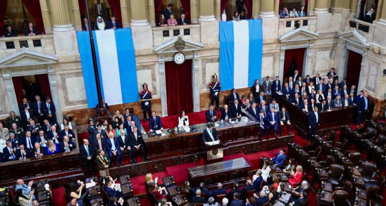 El presidente de Argentina, Javier Milei, de pie en el centro del Congreso, pronuncia un discurso durante la apertura de sesiones legislativas de 2025. A su alrededor, legisladores, funcionarios y autoridades, prestan atención a sus palabras. En el recinto destacan sus cortinas rojas y las banderas argentinas colgadas en las paredes, enmarcando el acto. Talvi