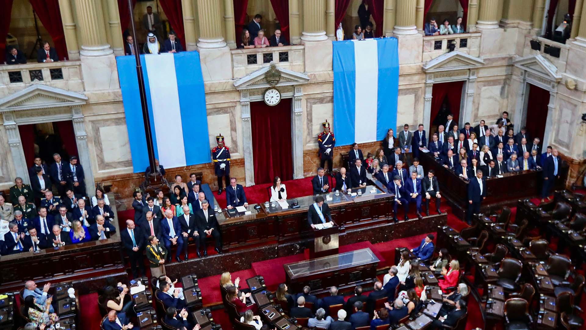 The President of Argentina, Javier Milei, stands at the centre of Congress, delivering a speech during the opening of the 2025 legislative session. Surrounding him, legislators, officials, and authorities listen attentively to his words. The chamber is distinguished by its red curtains and Argentine flags hanging on the walls, framing the event. Milei plan