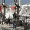 Descarga de gas natural licuado (GNL) en el puerto de Fos-sur-Mer, en Francia. Un buque metanero especializado en el transporte de GNL está atracado en la terminal, con los brazos de carga conectados para la transferencia del gas. Tres trabajadores, equipados con equipo de protección, supervisan la transferencia de gas desde una de las líneas hacia las instalaciones. El entorno es industrial, con estructuras metálicas, tuberías y válvulas visibles a la luz del día. Dominio energético