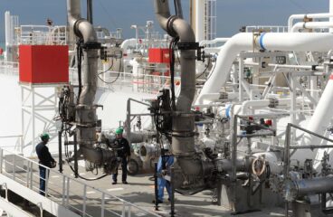 Descarga de gas natural licuado (GNL) en el puerto de Fos-sur-Mer, en Francia. Un buque metanero especializado en el transporte de GNL está atracado en la terminal, con los brazos de carga conectados para la transferencia del gas. Tres trabajadores, equipados con equipo de protección, supervisan la transferencia de gas desde una de las líneas hacia las instalaciones. El entorno es industrial, con estructuras metálicas, tuberías y válvulas visibles a la luz del día. Dominio energético