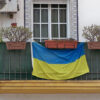 Balcón con barandilla verde y varias macetas, donde cuelga una bandera de Ucrania en Málaga (España). La ventana tiene un marco blanco con azulejos decorativos alrededor. A la izquierda, un aire acondicionado está instalado en la pared.