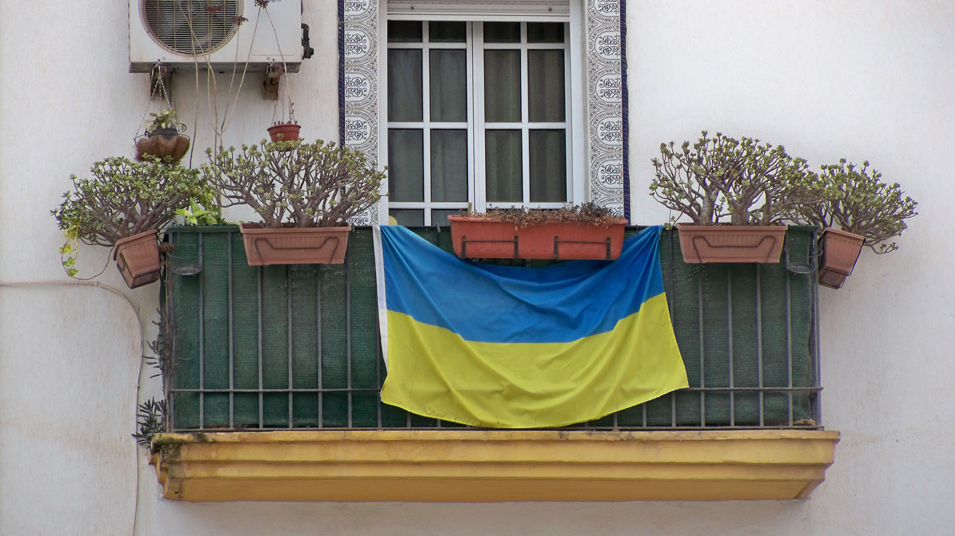 Balcón con barandilla verde y varias macetas, donde cuelga una bandera de Ucrania en Málaga (España). La ventana tiene un marco blanco con azulejos decorativos alrededor. A la izquierda, un aire acondicionado está instalado en la pared.