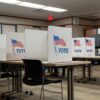 Cabinas de votación en la Biblioteca Pública de Aitkin, Minnesota (EEUU), durante las elecciones del 5 de noviembre de 2024. Varias cabinas blancas con la palabra "VOTE" y una bandera de Estados Unidos impresas están alineadas sobre mesas, brindando privacidad a los votantes. Algunas personas están sentadas mientras emiten su voto, aunque sus rostros no son visibles. En el fondo, hay máquinas de votación y una señal de salida con una flecha roja en la pared. La sala cuenta con iluminación fluorescente, paredes de color beige y un suelo alfombrado. Minorías