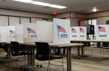 Cabinas de votación en la Biblioteca Pública de Aitkin, Minnesota (EEUU), durante las elecciones del 5 de noviembre de 2024. Varias cabinas blancas con la palabra "VOTE" y una bandera de Estados Unidos impresas están alineadas sobre mesas, brindando privacidad a los votantes. Algunas personas están sentadas mientras emiten su voto, aunque sus rostros no son visibles. En el fondo, hay máquinas de votación y una señal de salida con una flecha roja en la pared. La sala cuenta con iluminación fluorescente, paredes de color beige y un suelo alfombrado. Minorías