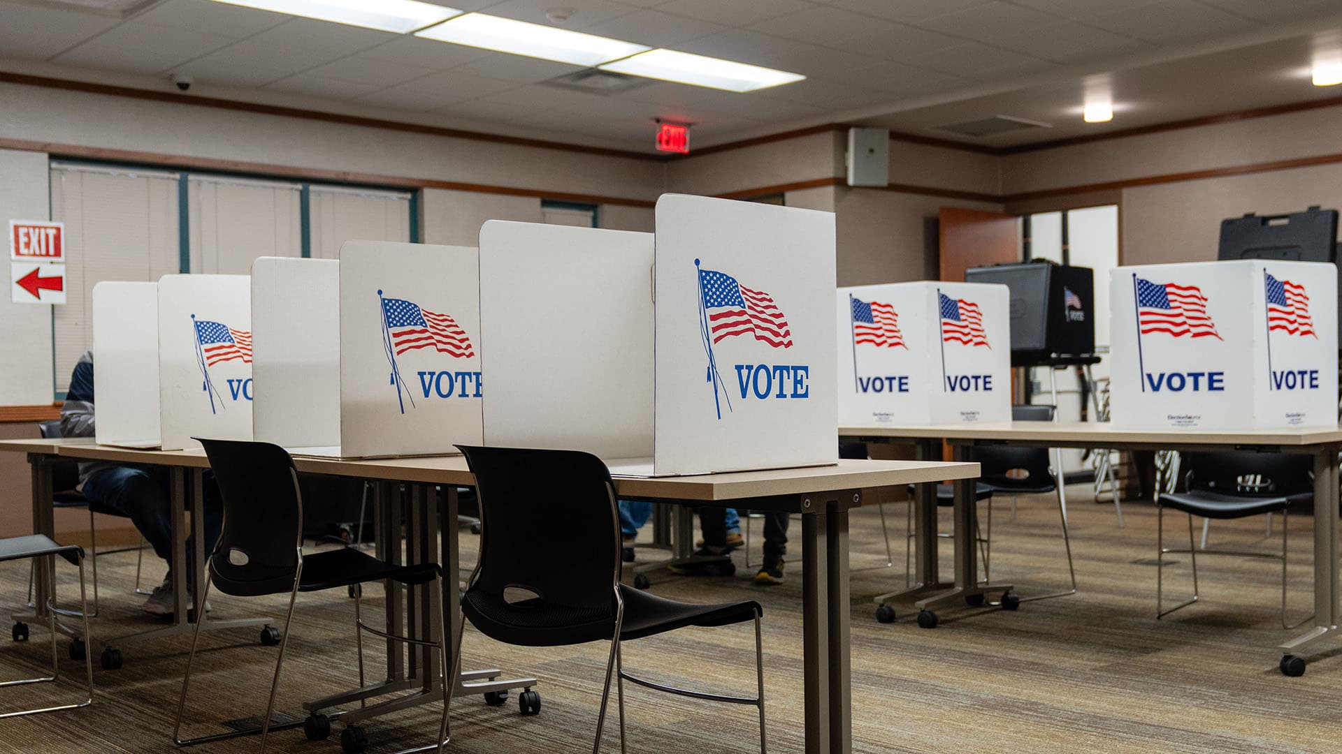 Cabinas de votación en la Biblioteca Pública de Aitkin, Minnesota (EEUU), durante las elecciones del 5 de noviembre de 2024. Varias cabinas blancas con la palabra "VOTE" y una bandera de Estados Unidos impresas están alineadas sobre mesas, brindando privacidad a los votantes. Algunas personas están sentadas mientras emiten su voto, aunque sus rostros no son visibles. En el fondo, hay máquinas de votación y una señal de salida con una flecha roja en la pared. La sala cuenta con iluminación fluorescente, paredes de color beige y un suelo alfombrado. Minorías