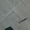 Aerial view of a basketball court with worn-out lines and a cracked surface. Two people are playing on the court, casting long shadows under the sunlight. Education