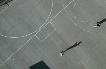 Aerial view of a basketball court with worn-out lines and a cracked surface. Two people are playing on the court, casting long shadows under the sunlight. Education