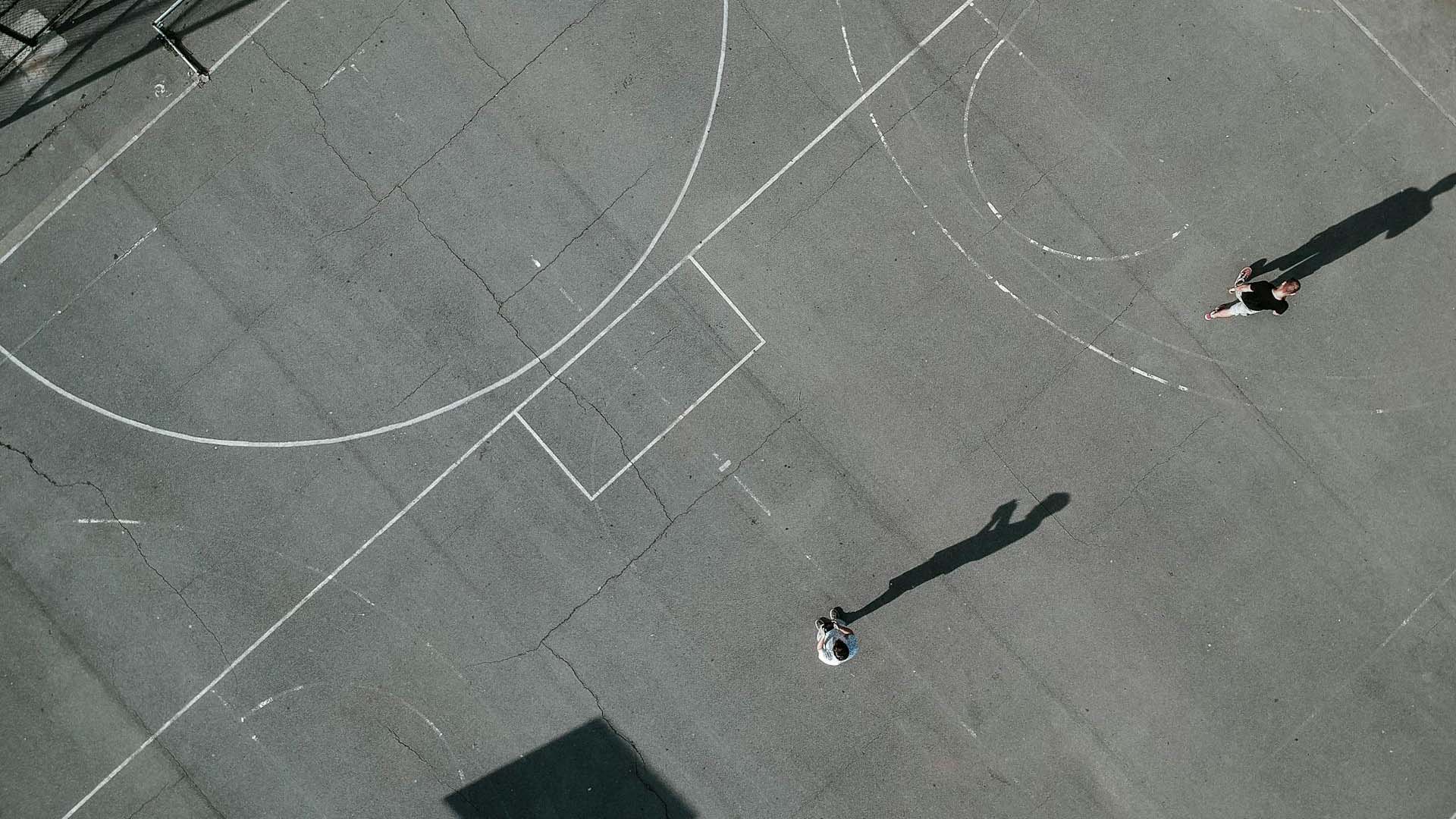 Aerial view of a basketball court with worn-out lines and a cracked surface. Two people are playing on the court, casting long shadows under the sunlight. Education