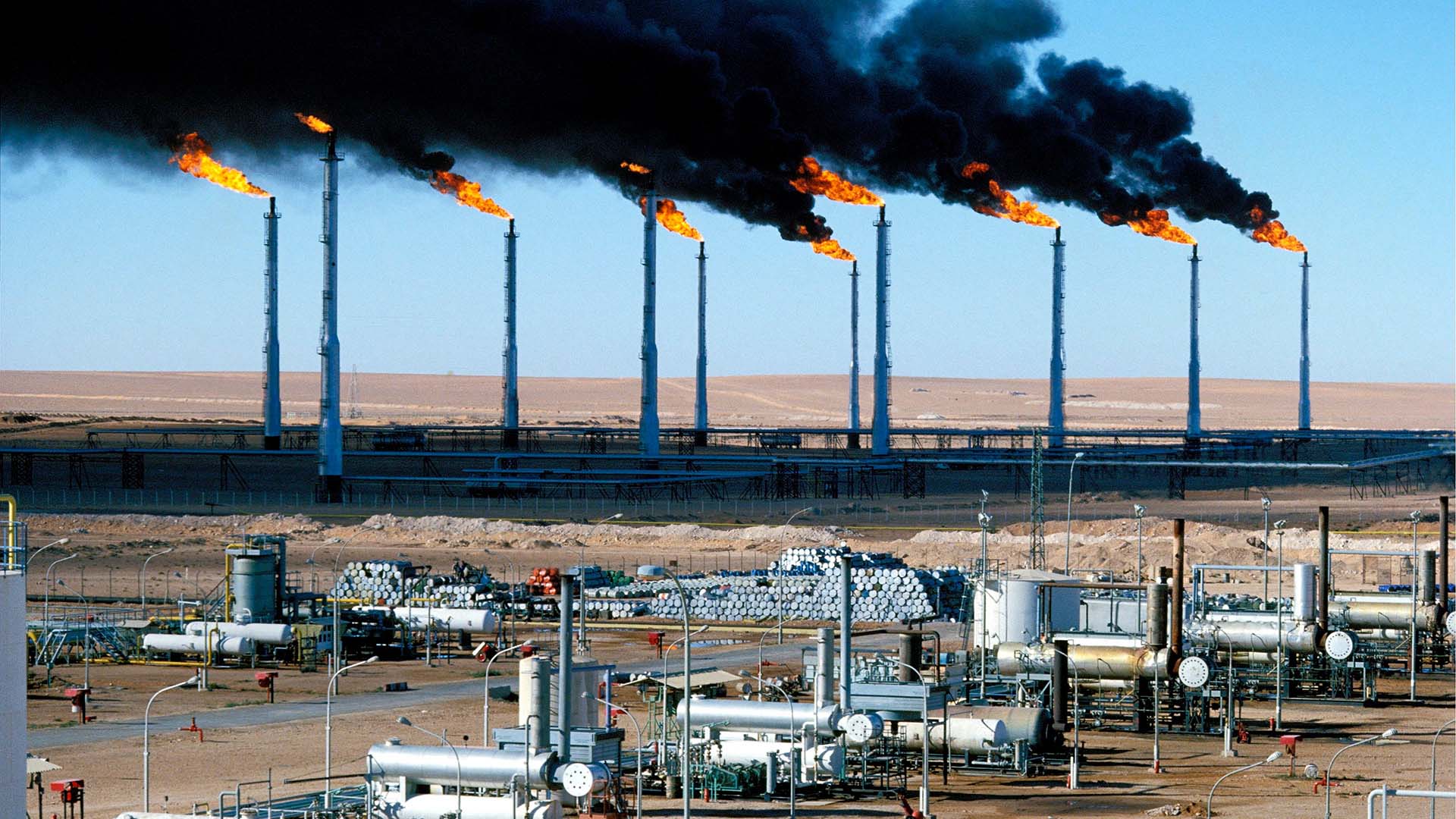 Hassi R’Mel, el mayor campo y hub de gas en Argelia, con múltiples antorchas de gas quemando y emitiendo humo negro sobre una vasta instalación industrial en el desierto. Gas