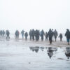 Un grupo de personas migrantes camina por la playa de Petit-Fort-Philippe, en Gravelines, Francia, el 17 de enero de 2025, mientras se preparan para partir en barco hacia el Reino Unido. Las personas, de espaldas a la cámara, avanzan hacia la embarcación en un día gris, con niebla y cielo nublado. Europa